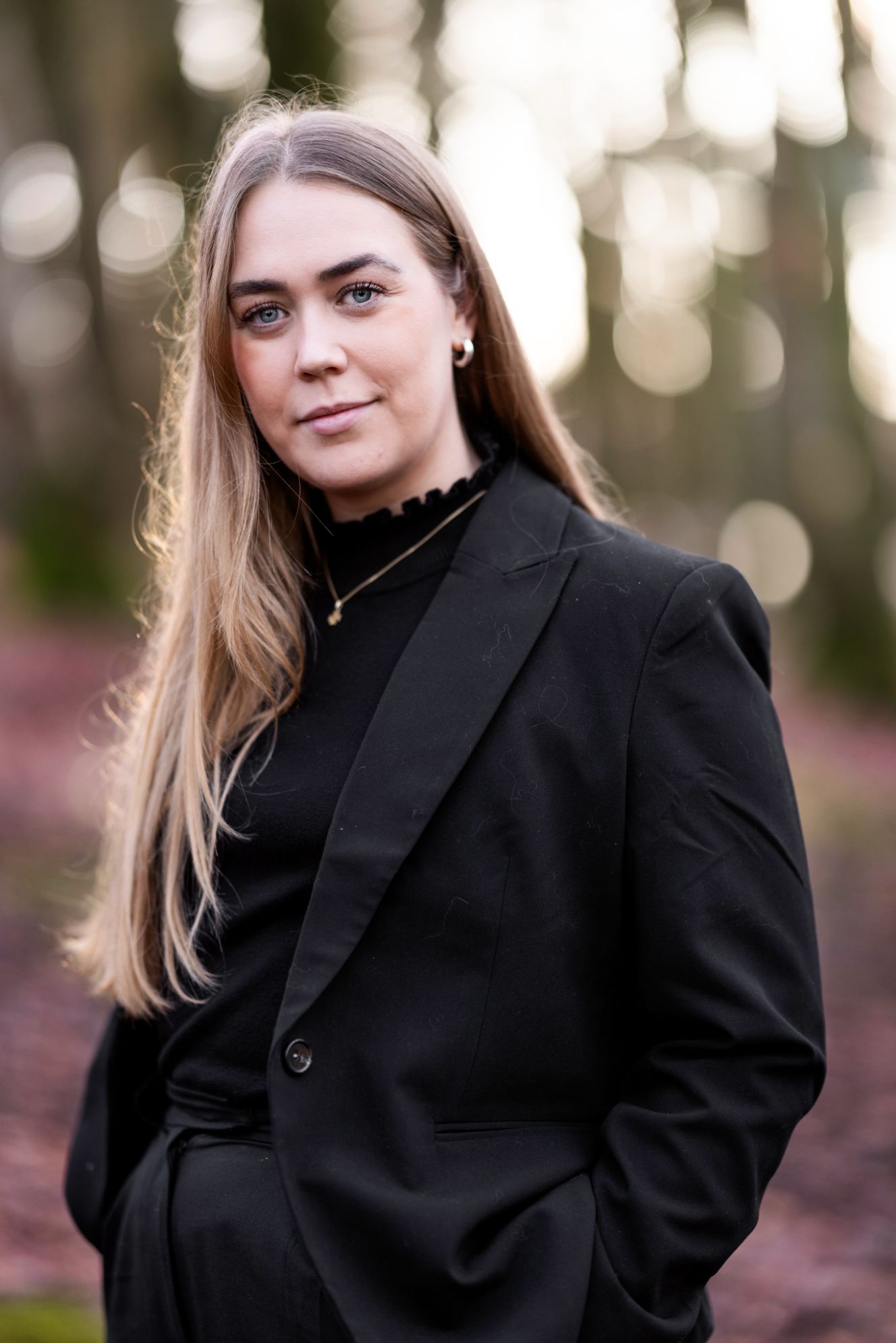 En kvinne i dress ser alvorlig inn i kameraet i en skog.