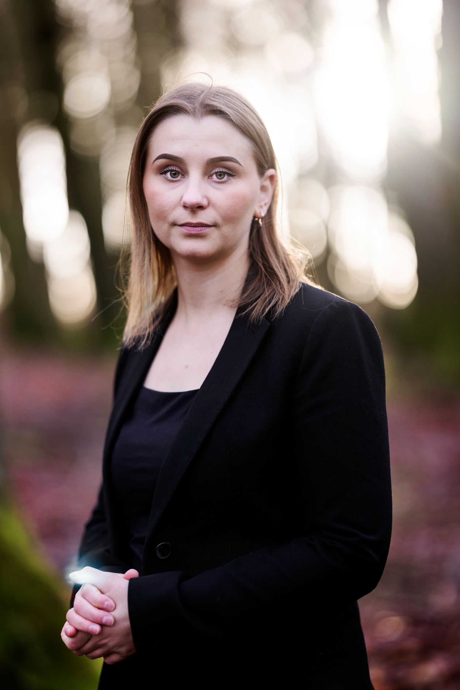 En kvinne i dress med foldede hender står i en skog.