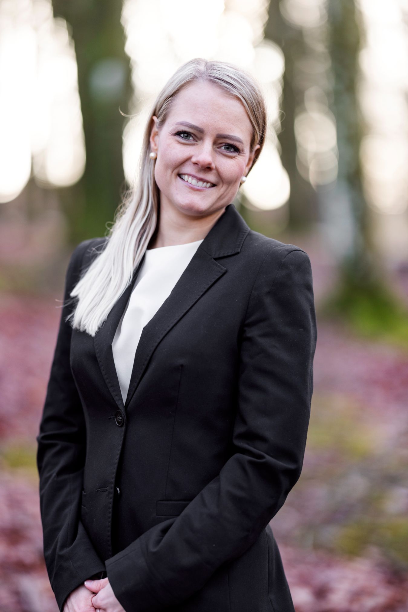 En smilende kvinne i dress står i en skog.